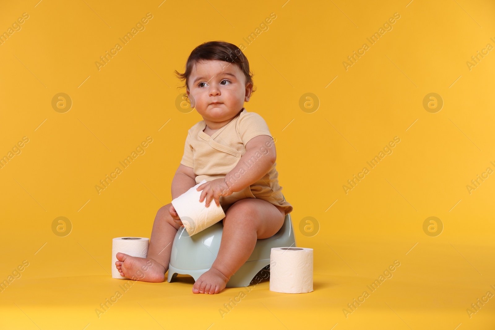 Cute little baby sitting on potty and rolls of toilet paper against orange background Photo of Cute little baby sitting on potty and rolls of toilet paper against orange background
