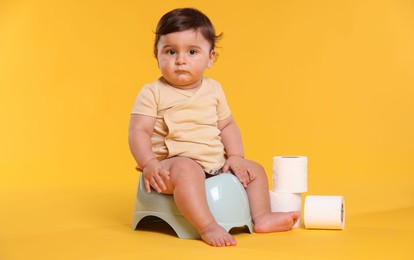 Photo of Cute little baby sitting on potty and rolls of toilet paper against orange background