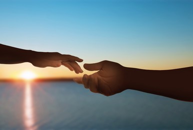 Image of Man and woman reaching to each other against seascape at sunset, closeup of hands