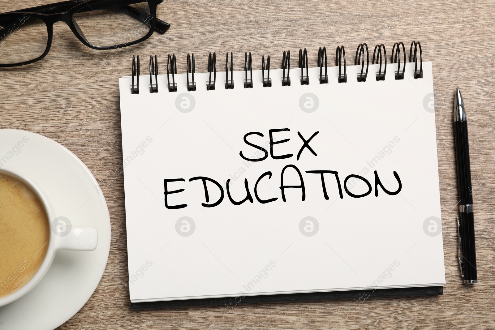 Sex Education inscription in notebook, pen, glasses and coffee on wooden table, top view Image of Sex Education inscription in notebook, pen, glasses and coffee on wooden table, top view