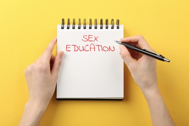 Image of Woman writing Sex Education inscription in notebook on orange background, top view