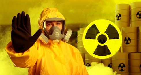 Image of Radioactive hazard. Man in hazmat suit and gas mask blocking way to barrels with warning signs behind him