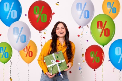 Image of Hot sale, special offer. Happy woman holding gift box on white background. Colorful balloons with percent signs around her