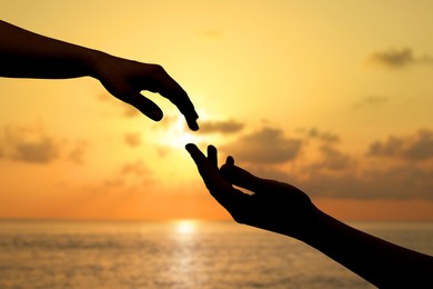 Image of Man and woman reaching to each other against seascape at sunset, closeup of hands
