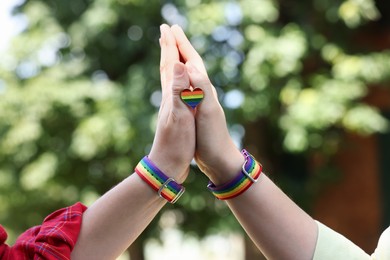 Photo of LGBT concept. Women in rainbow wristbands with heart shaped pin holding hands outdoors, closeup