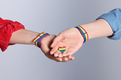 Photo of LGBT concept. Women in rainbow wristbands with heart shaped pin holding hands on light grey background, closeup