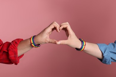 Photo of LGBT concept. Women in rainbow wristbands making heart shape on pink background, closeup
