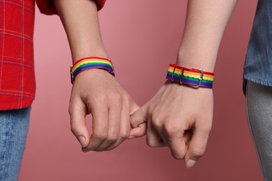 Photo of LGBT concept. Women in rainbow wristbands holding hands on pink background, closeup