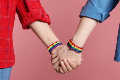 Photo of LGBT concept. Women in rainbow wristbands holding hands on pink background, closeup