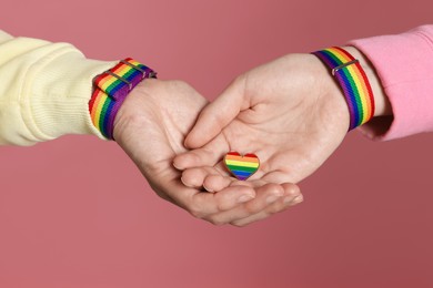 Photo of LGBT concept. Women in rainbow wristbands with heart shaped pin holding hands on pink background, closeup