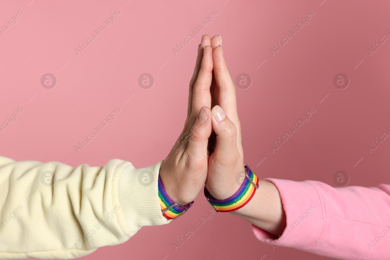 Photo of LGBT concept. Women in rainbow wristbands holding hands on pink background, closeup