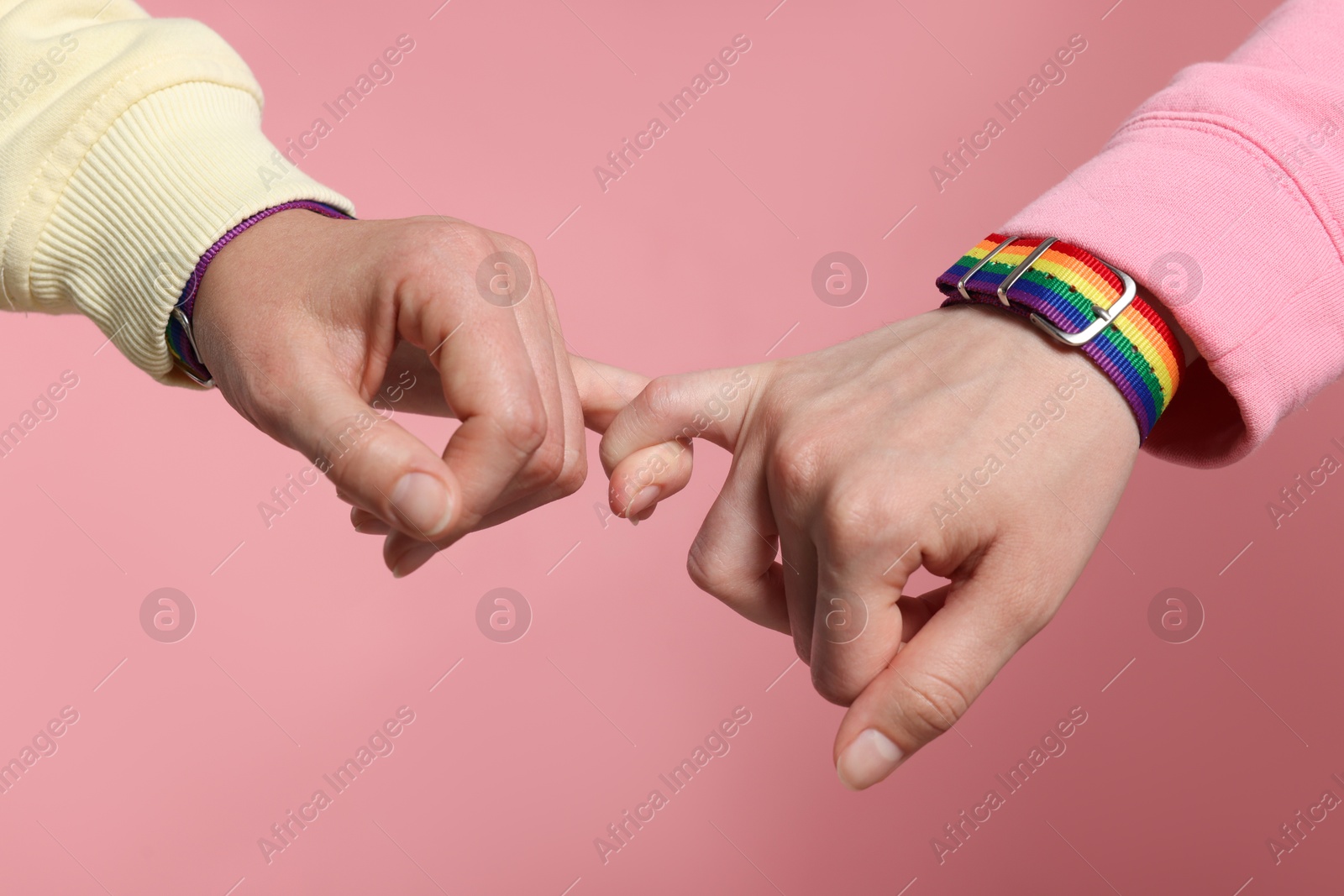 Photo of LGBT concept. Women in rainbow wristbands holding hands on pink background, closeup