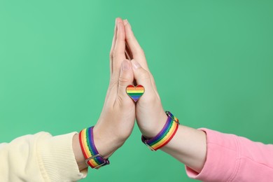Photo of LGBT concept. Women in rainbow wristbands with heart shaped pin holding hands on green background, closeup