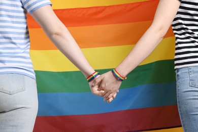 Photo of LGBT concept. Women in rainbow wristbands holding hands against flag, closeup