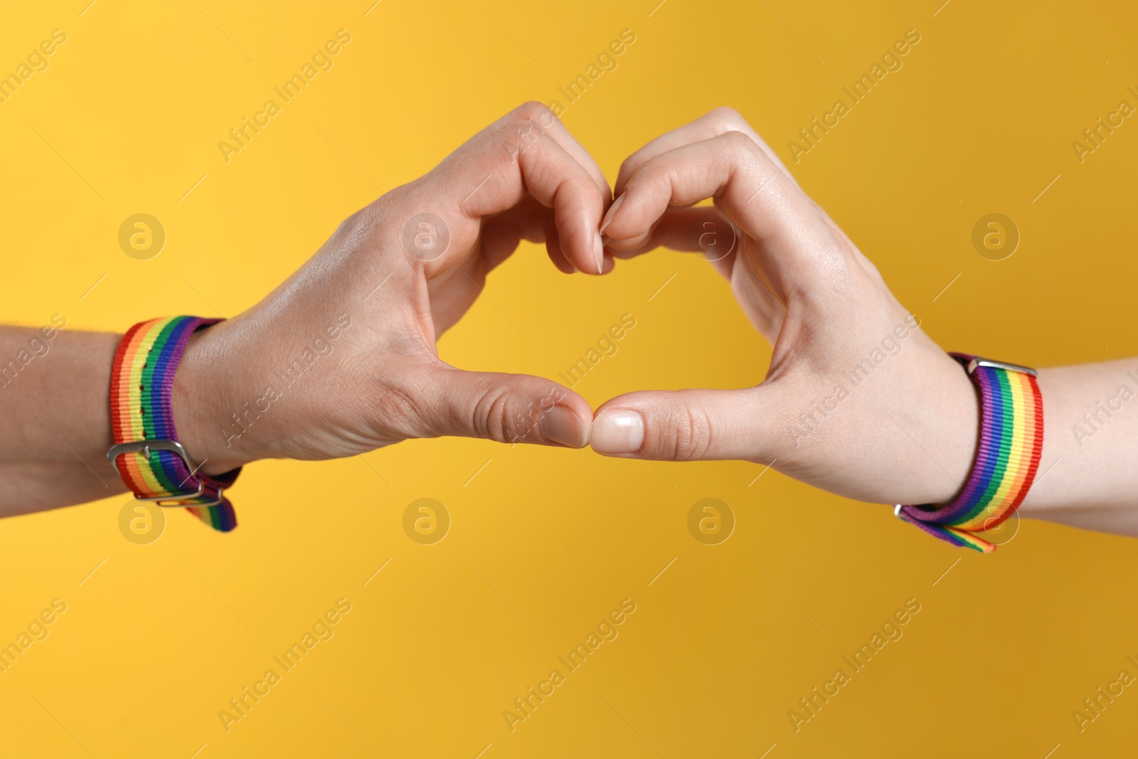 Photo of LGBT concept. Women in rainbow wristbands making heart shape on yellow background, closeup