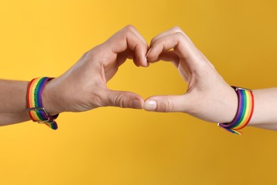 Photo of LGBT concept. Women in rainbow wristbands making heart shape on yellow background, closeup