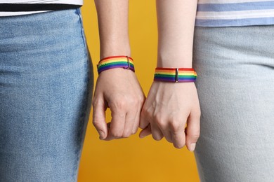 Photo of LGBT concept. Women in rainbow wristbands holding hands on yellow background, closeup