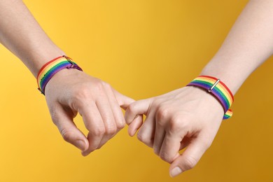 Photo of LGBT concept. Women in rainbow wristbands holding hands on yellow background, closeup