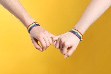 Photo of LGBT concept. Women in rainbow wristbands holding hands on yellow background, closeup