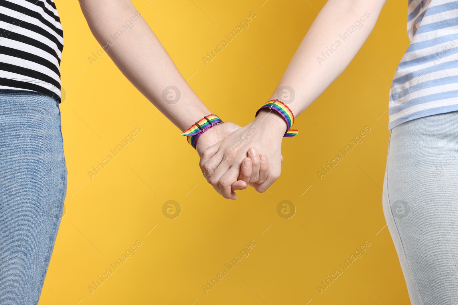 Photo of LGBT concept. Women in rainbow wristbands holding hands on yellow background, closeup