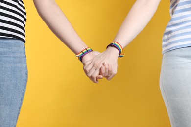 Photo of LGBT concept. Women in rainbow wristbands holding hands on yellow background, closeup