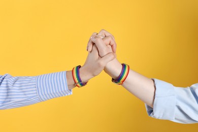 Photo of LGBT concept. Women in rainbow wristbands holding hands on yellow background, closeup