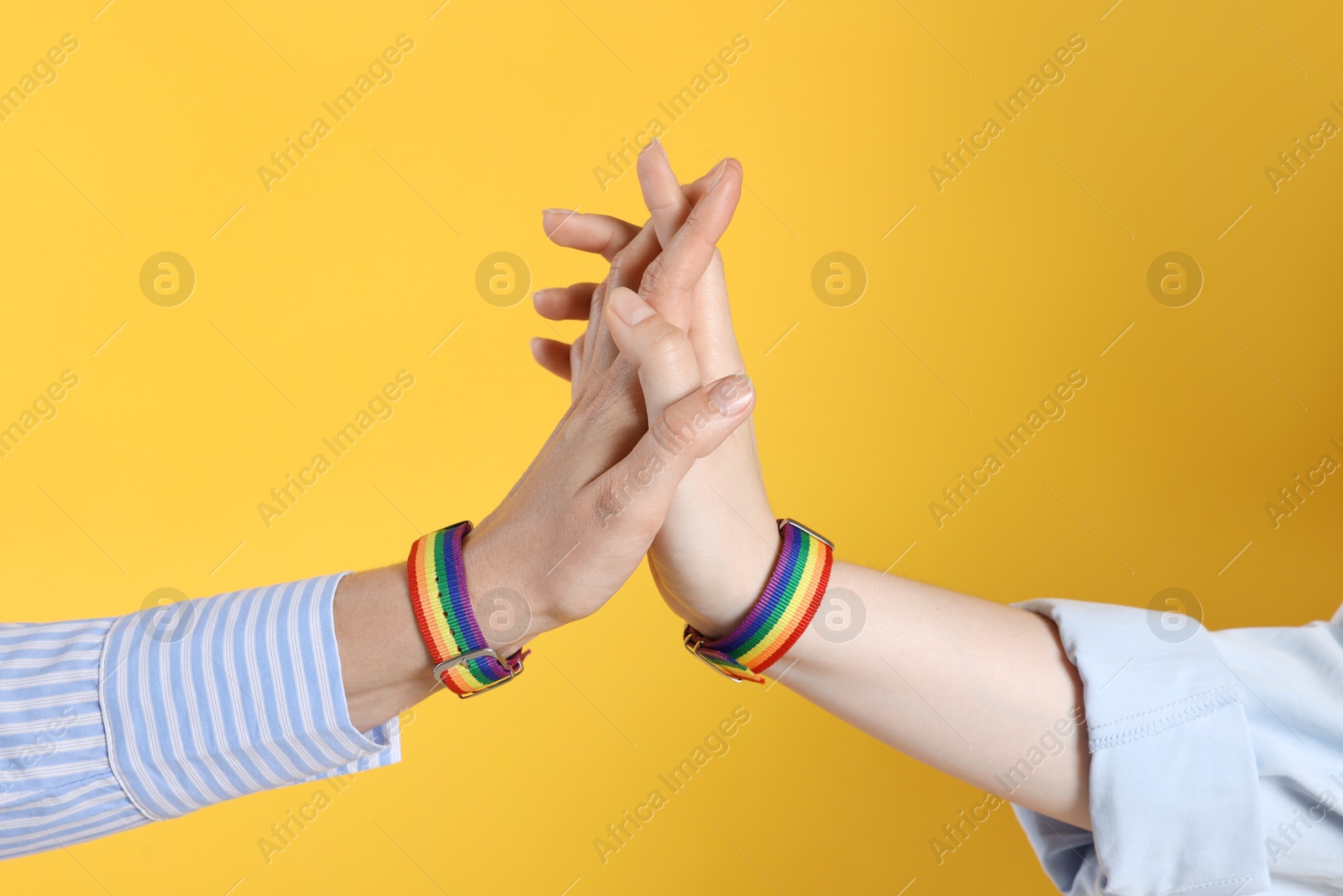 Photo of LGBT concept. Women in rainbow wristbands holding hands on yellow background, closeup