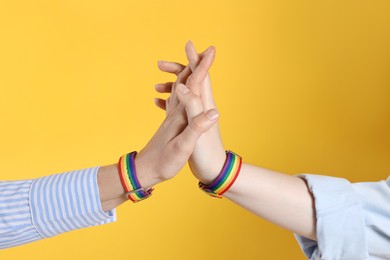 Photo of LGBT concept. Women in rainbow wristbands holding hands on yellow background, closeup