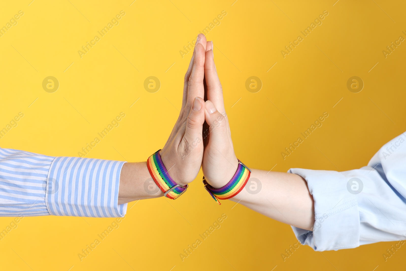 Photo of LGBT concept. Women in rainbow wristbands holding hands on yellow background, closeup