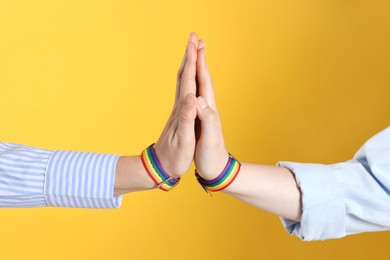 Photo of LGBT concept. Women in rainbow wristbands holding hands on yellow background, closeup