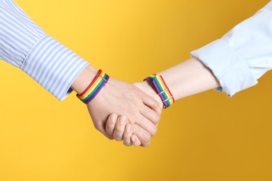 Photo of LGBT concept. Women in rainbow wristbands holding hands on yellow background, closeup