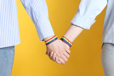 Photo of LGBT concept. Women in rainbow wristbands holding hands on yellow background, closeup