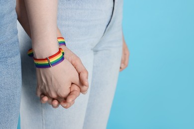 Photo of LGBT concept. Women in rainbow wristbands holding hands on light blue background, closeup. Space for text