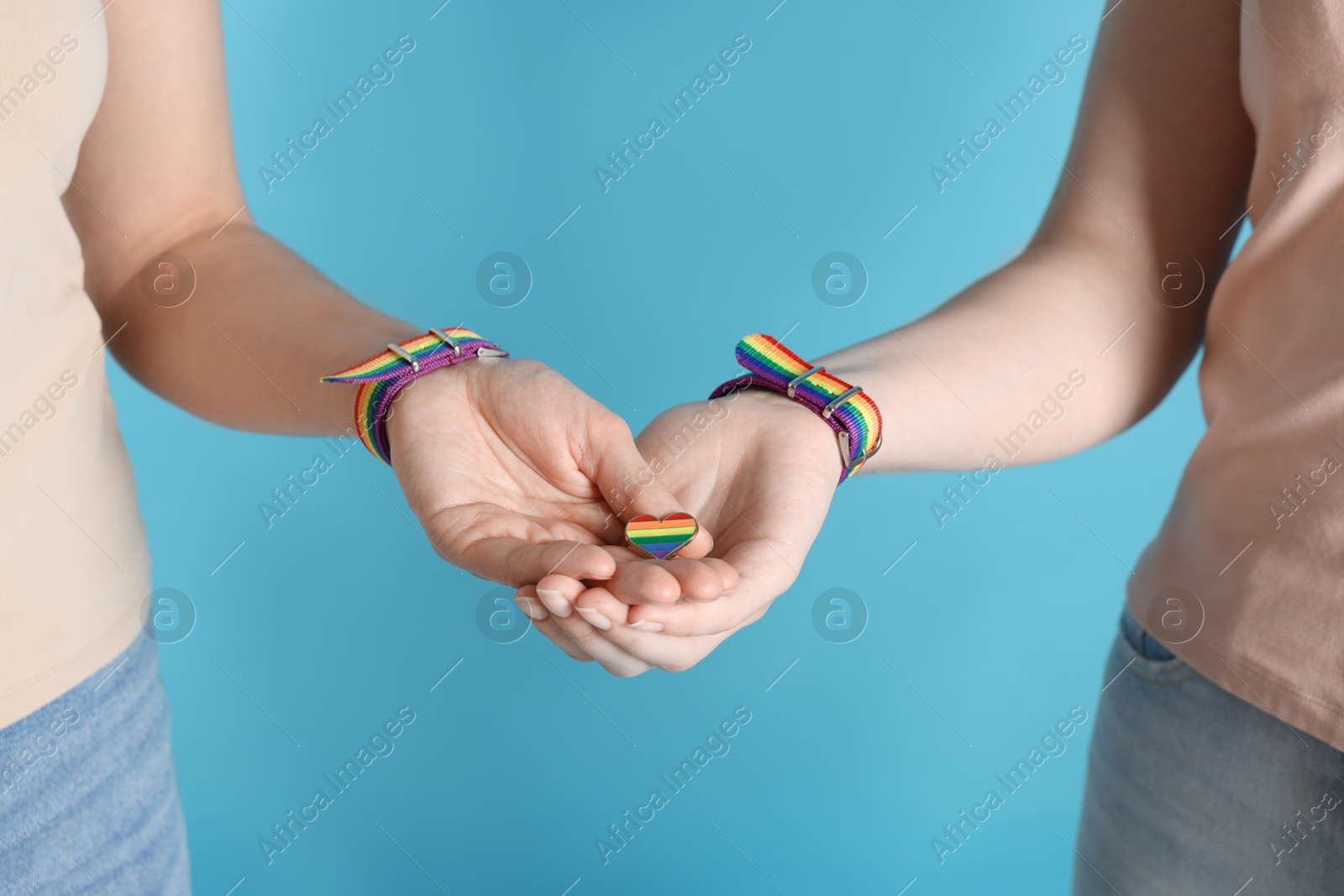LGBT concept. Women in rainbow wristbands with heart shaped pin holding hands on light blue background, closeup Photo of LGBT concept. Women in rainbow wristbands with heart shaped pin holding hands on light blue background, closeup