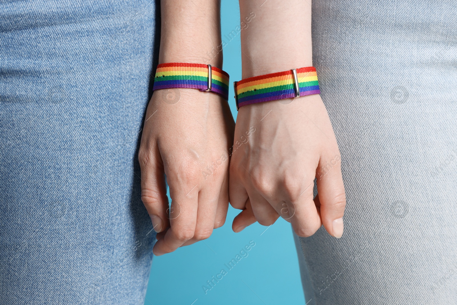 Photo of LGBT concept. Women in rainbow wristbands holding hands on light blue background, closeup