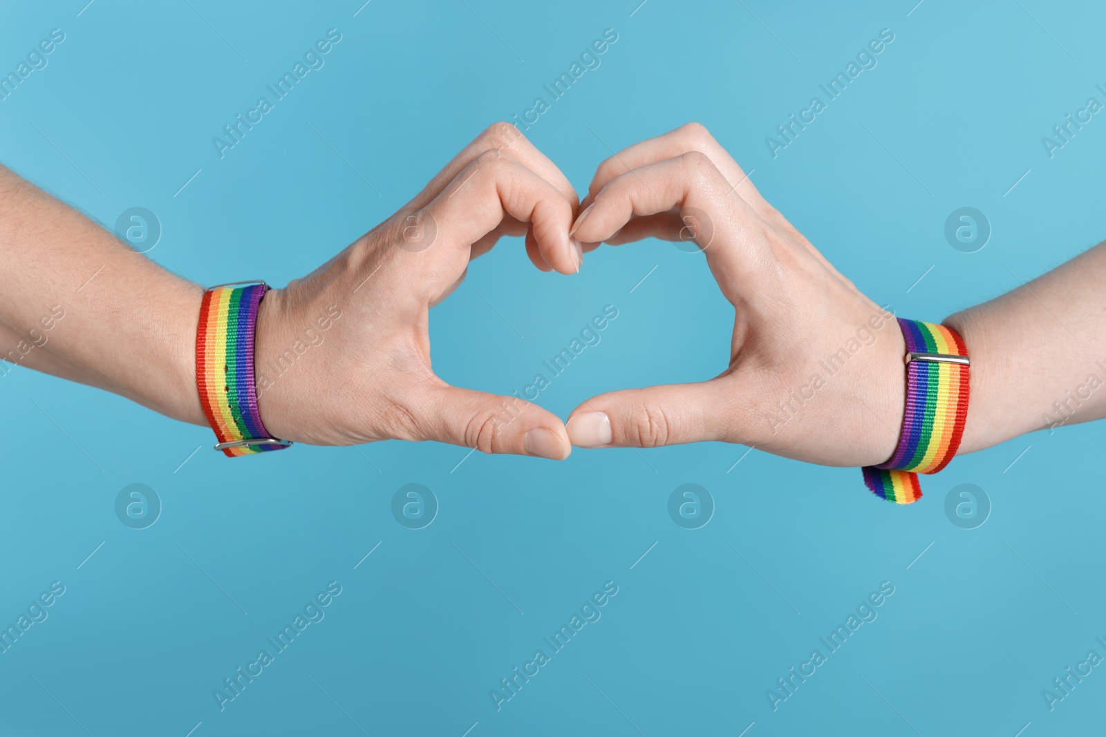 LGBT concept. Women in rainbow wristbands making heart shape on light blue background, closeup Photo of LGBT concept. Women in rainbow wristbands making heart shape on light blue background, closeup