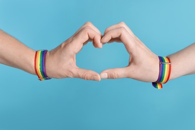 Photo of LGBT concept. Women in rainbow wristbands making heart shape on light blue background, closeup