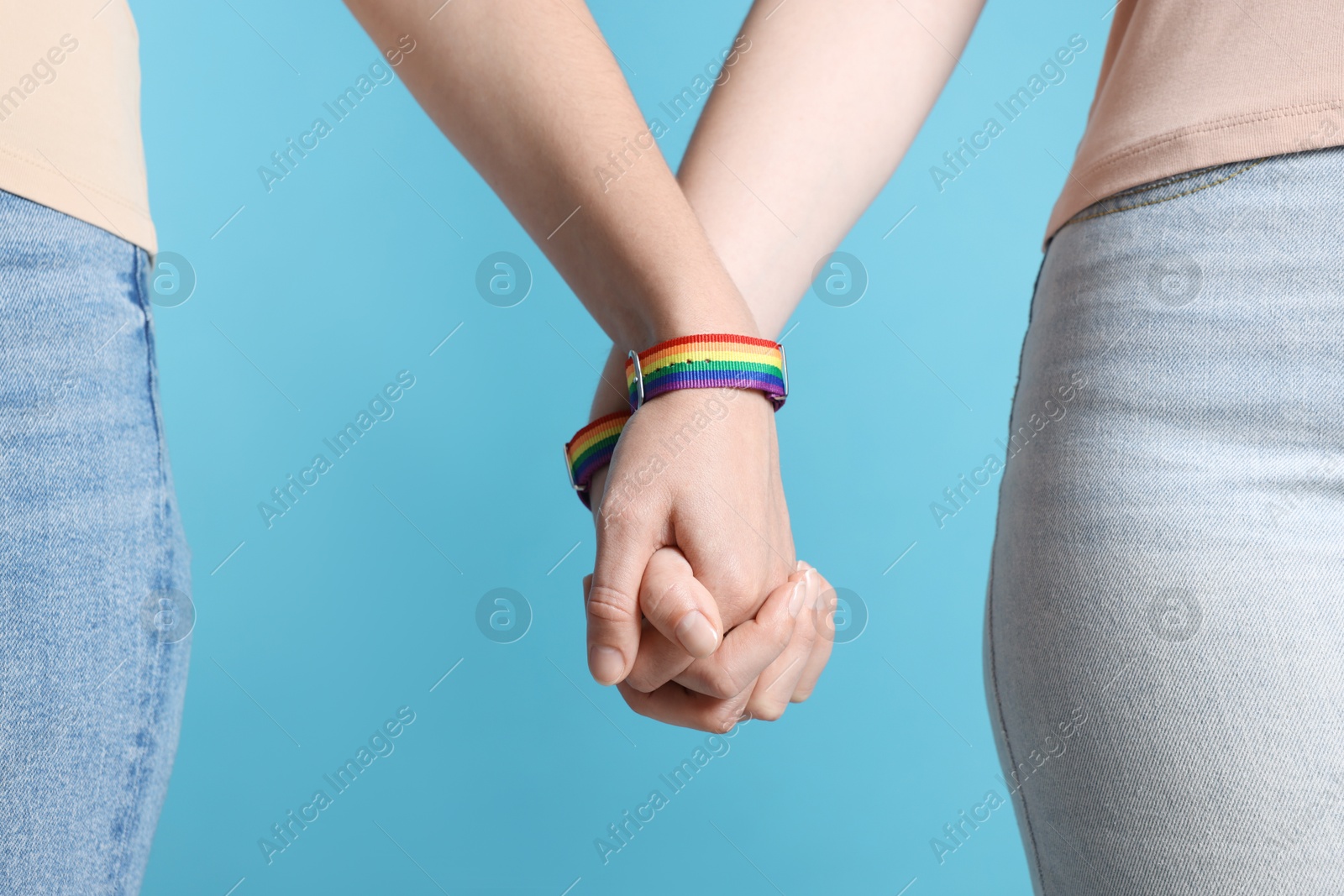 Photo of LGBT concept. Women in rainbow wristbands holding hands on light blue background, closeup