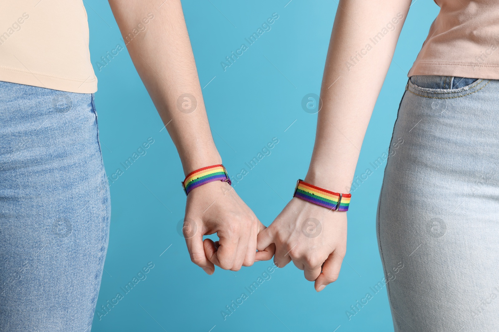 Photo of LGBT concept. Women in rainbow wristbands holding hands on light blue background, closeup