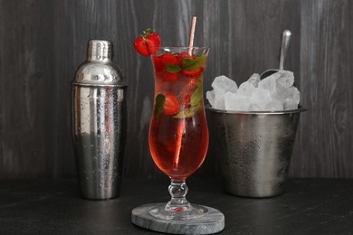 Photo of Refreshing strawberry cocktail in glass, metal shaker and ice in bucket on dark table