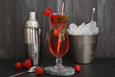 Photo of Refreshing strawberry cocktail in glass, metal shaker and ice in bucket on dark table