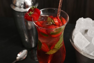 Photo of Refreshing strawberry cocktail in glass, metal shaker and ice in bucket on dark table, closeup