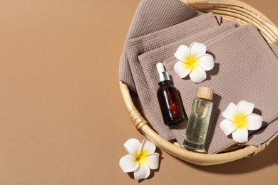 Photo of Clean waffle towels, bottles of cosmetic products and plumeria flowers in wicker basket on light brown background, flat lay. Space for text