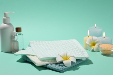 Photo of Folded waffle towels, plumeria flowers, burning candles and spa products on turquoise background, closeup
