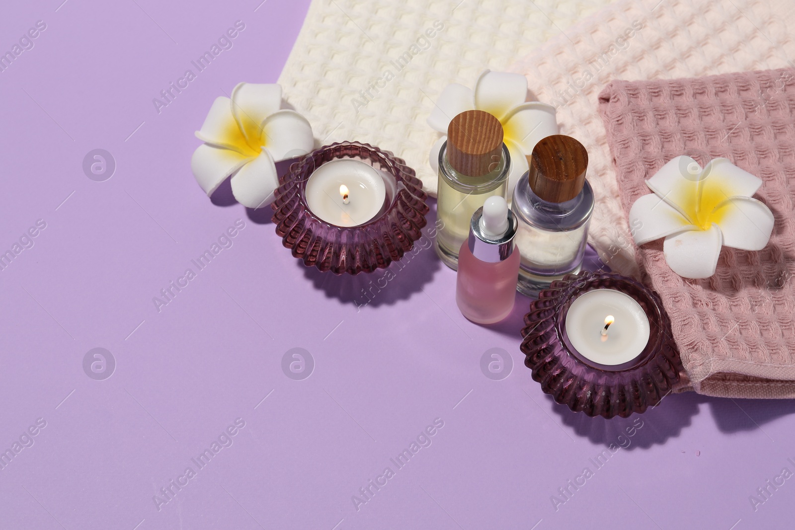 Photo of Folded waffle towels, burning candles, spa products and plumeria flowers on violet background. Space for text