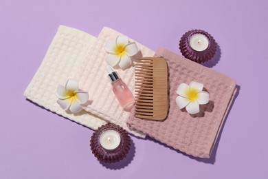 Photo of Folded waffle towels, burning candles, dropper and plumeria flowers on violet background, flat lay