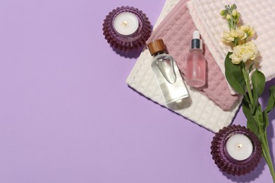 Photo of Folded waffle towels, burning candles, spa products and flowers on violet background, flat lay. Space for text
