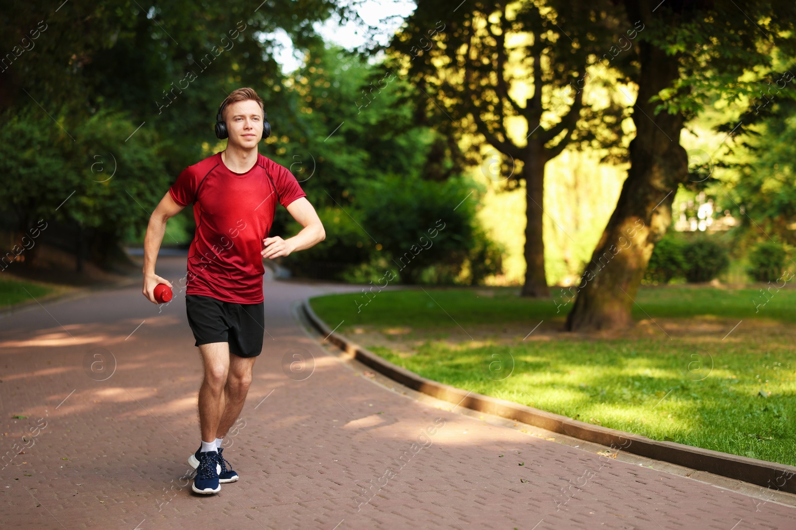 Photo of Smiling sportsman in headphones running with bottle outdoors. Morning exercise