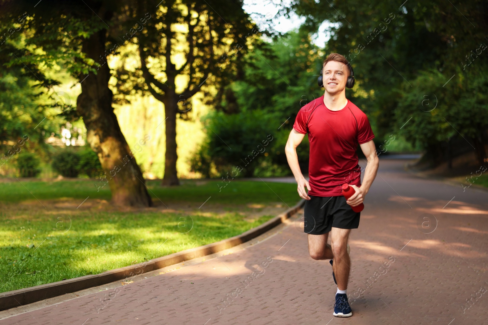 Photo of Smiling sportsman in headphones running with bottle outdoors. Morning exercise