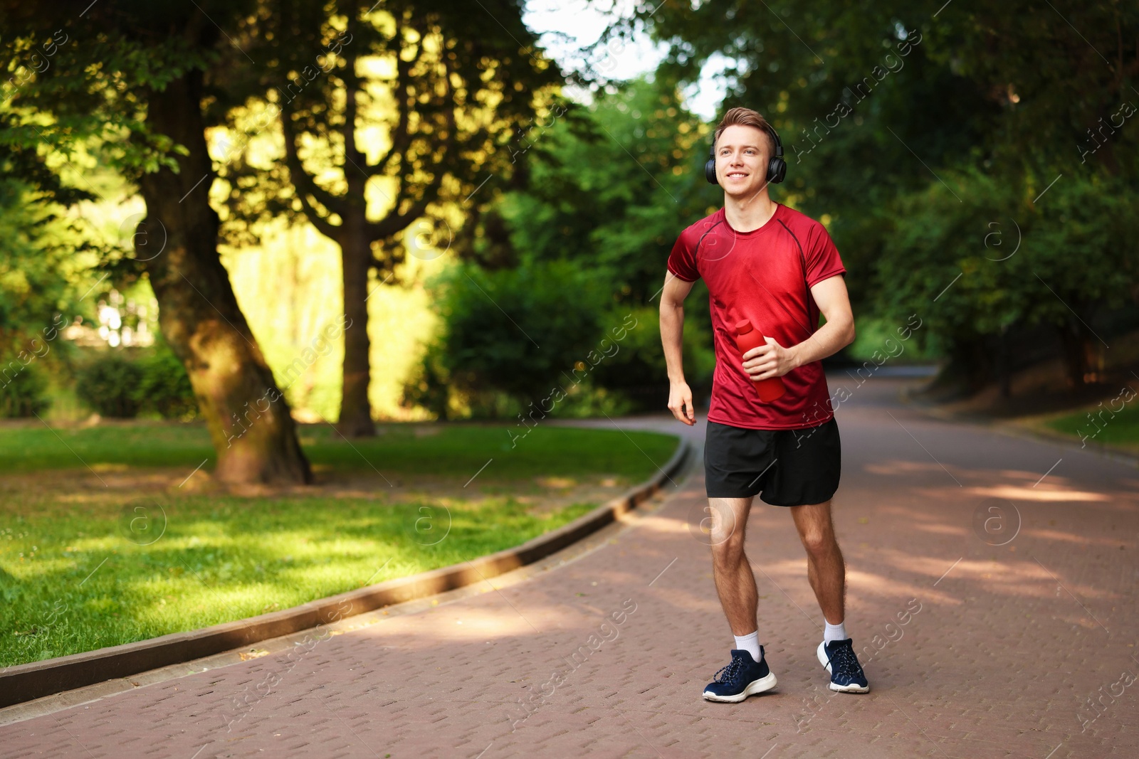 Photo of Smiling sportsman in headphones running with bottle outdoors. Morning exercise